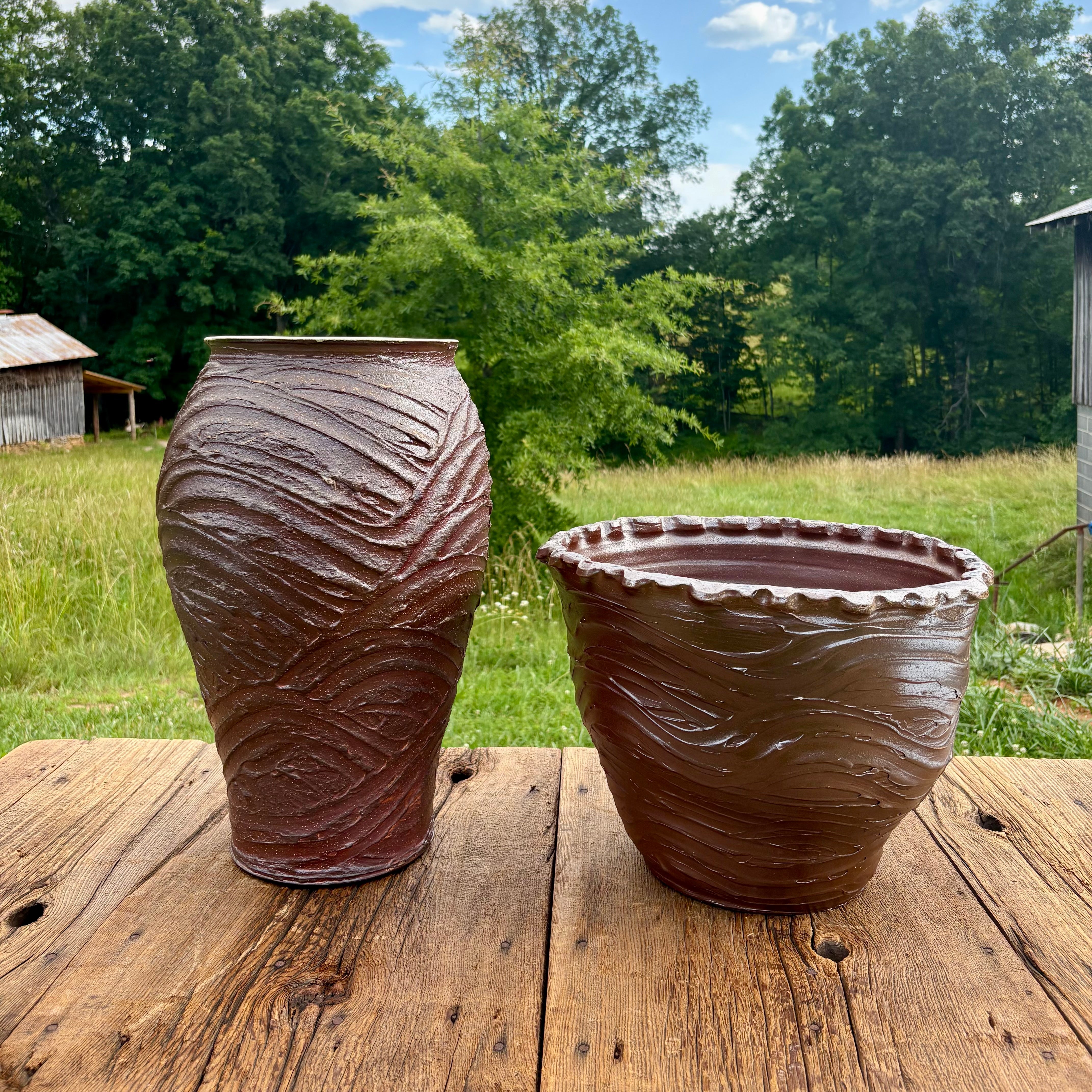Two large shiny brown pottery pots with a wave like pattern on a wooden table and rolling stoke county, NC hills in the background