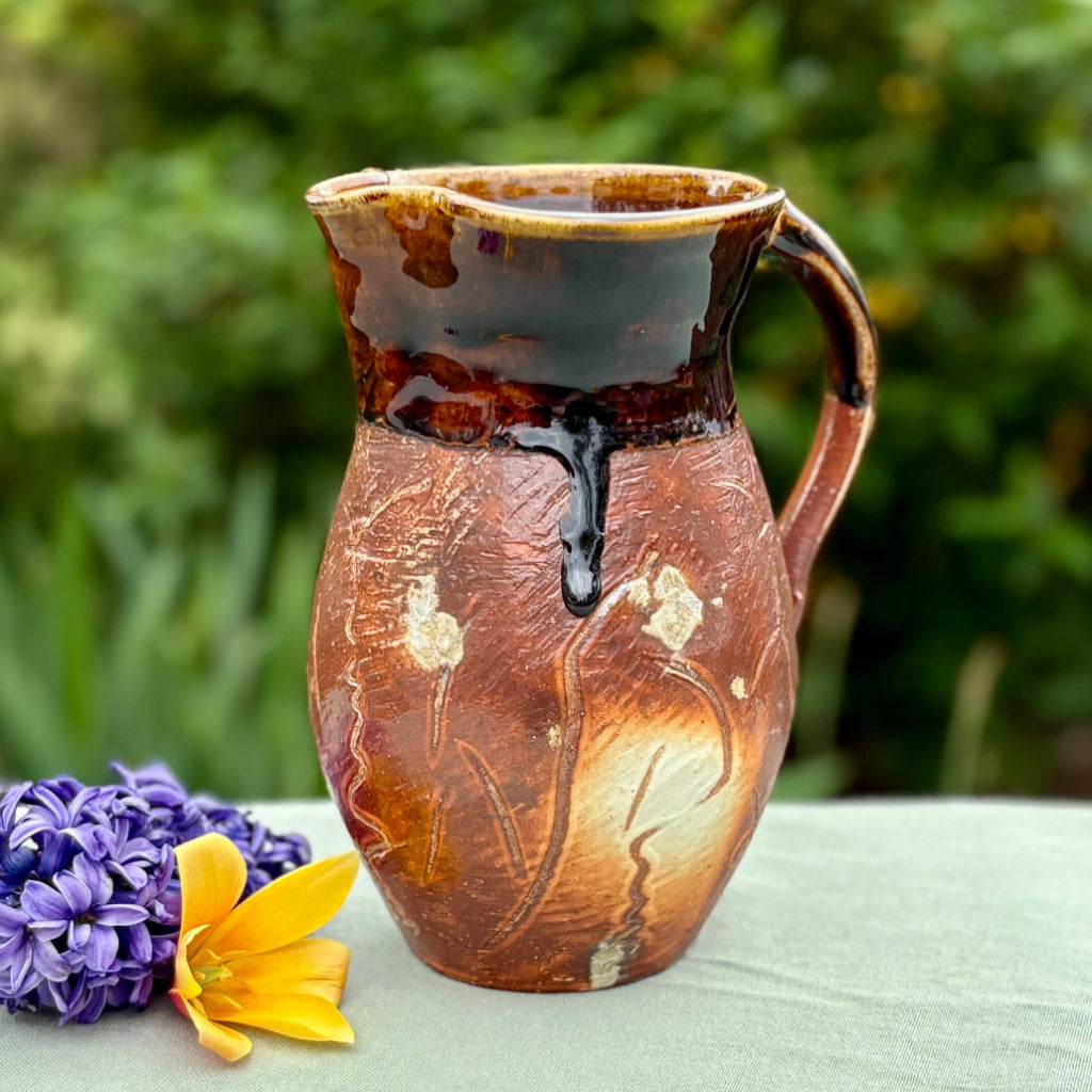 Handcrafted amber and chocolate brown wood-fired stoneware pitcher with pouring spout and handle on a green cloth with flowers and greenery in the background.
