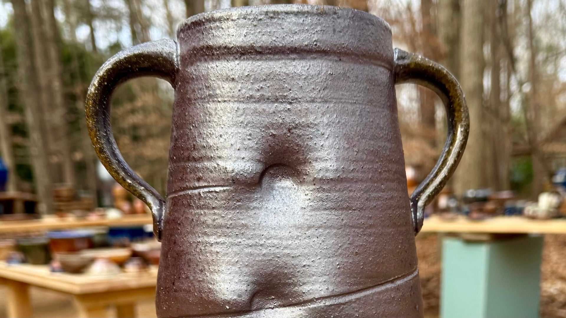 Brown pottery jug with two large handles with nature and pottery on tables in the background.