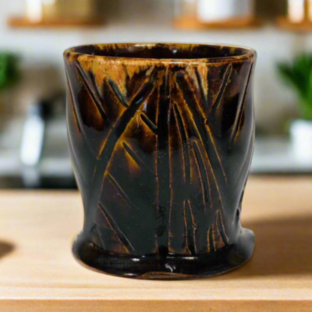 Handmade tenmoku cup with carved geometric pattern and amber rim, artisan ceramic North Carolina pottery on a wooden countertop with plants and shelves in the background.