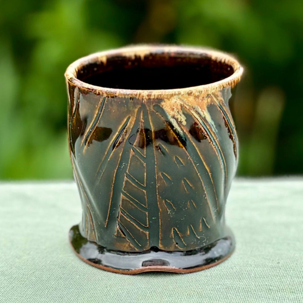 Handmade tenmoku cup with carved geometric pattern and amber rim, artisan ceramic North Carolina pottery on a green cloth with greenery in the background.
