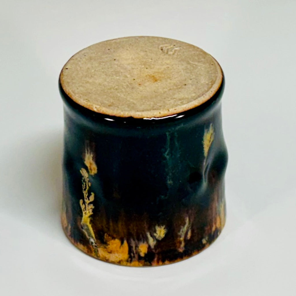 Small ceramic cup with a natural clay bottom and amber and beige runs on the exterior