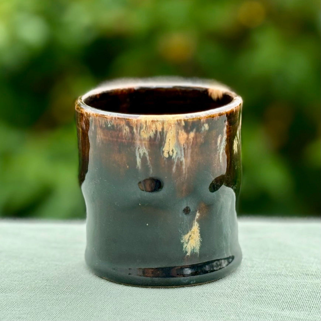 A lovely brown glazed cup with a beige and amber runs on a green cloth with greenery.
