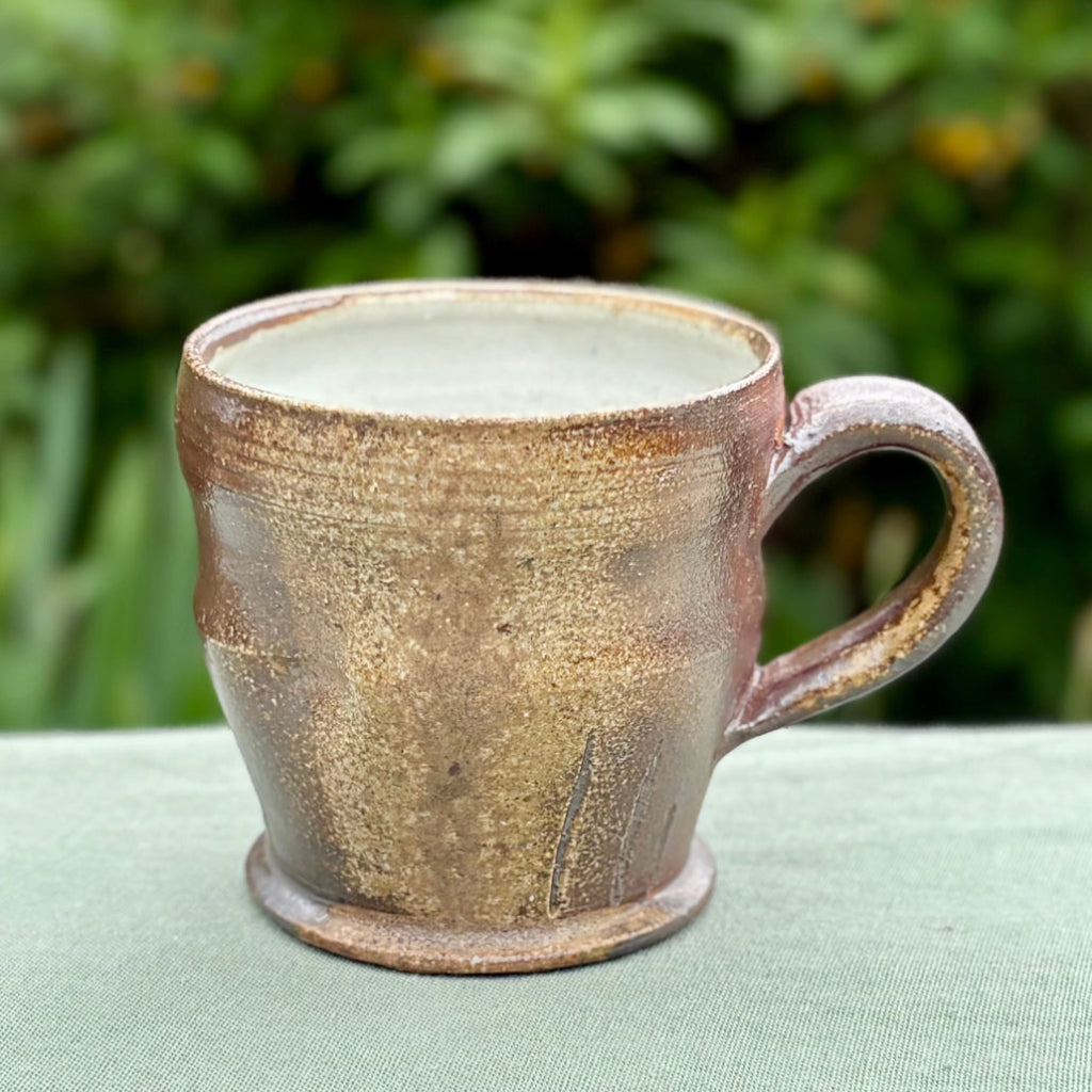 Handcrafted wood-fired brown stoneware mug with white interior and curved handle from Danbury pottery on a green cloth with greenery in the background.