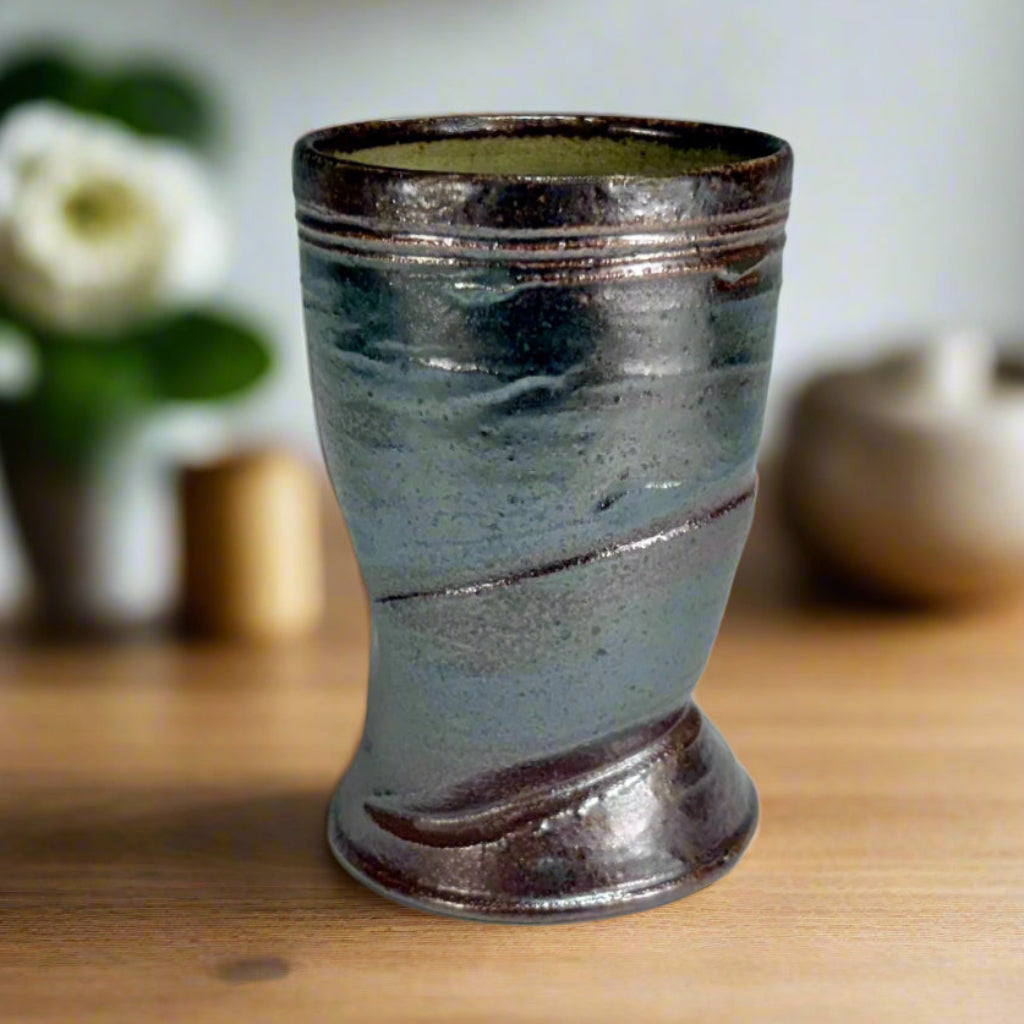 handcrafted wood-fired pottery cup with green-dark brown exterior on wooden counter with flowers in the background.