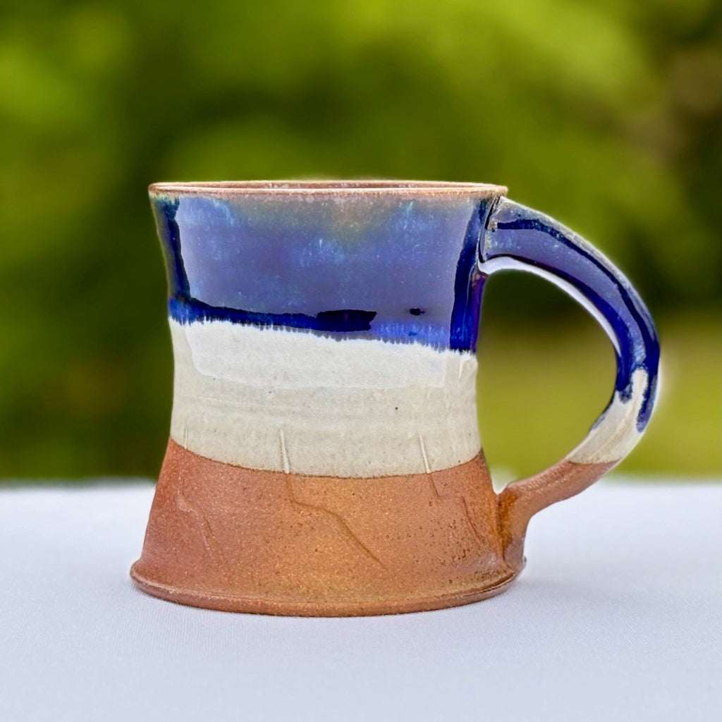 Ceramic mug with blue handle and brown base on a white surface with green blurred background