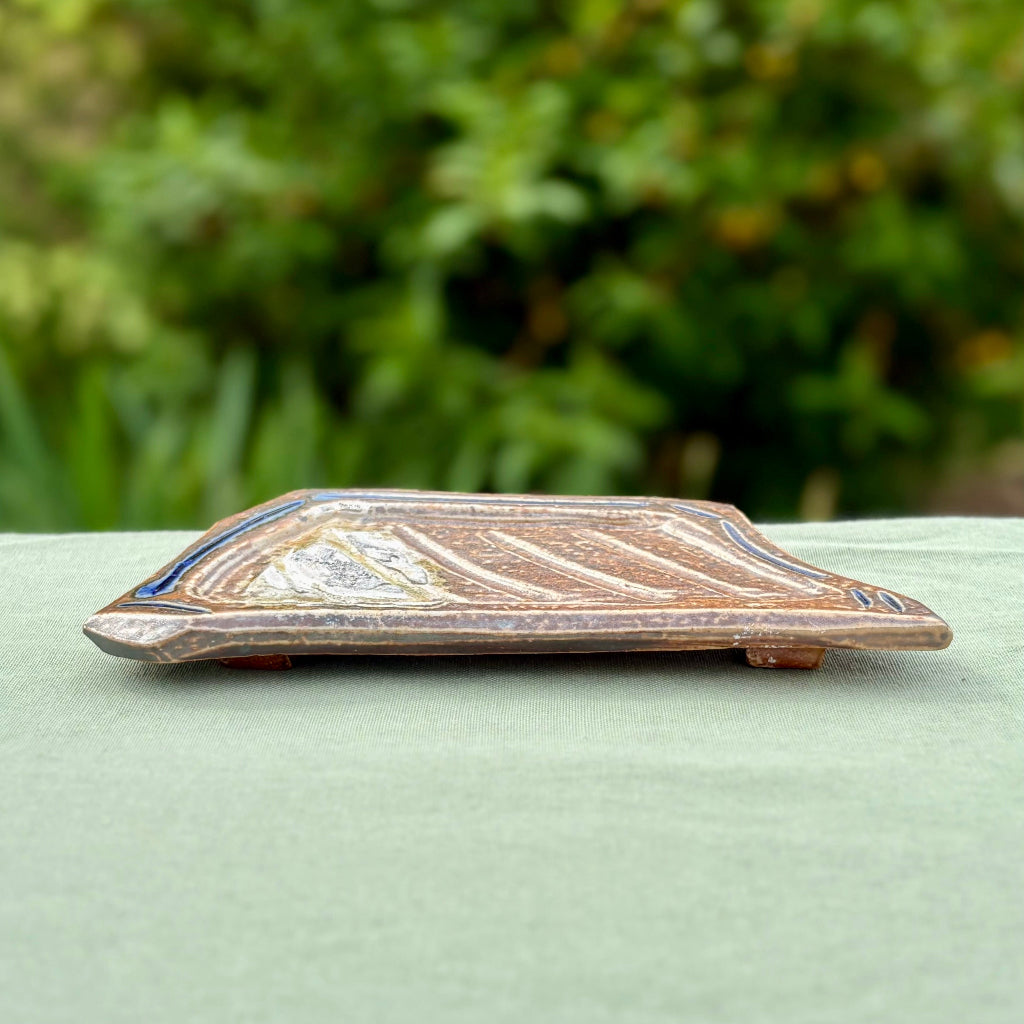 beige and blue pottery soap dish with a traditional elongated shape, displayed against a blurred greenery background