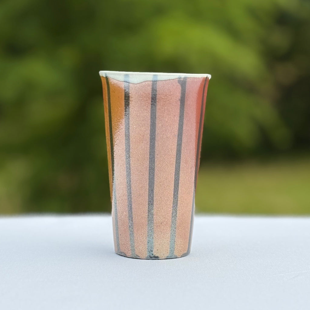 Porcelain tumbler with vertical black stripes that contrast with the soft peach and orange color of the unglazed cup