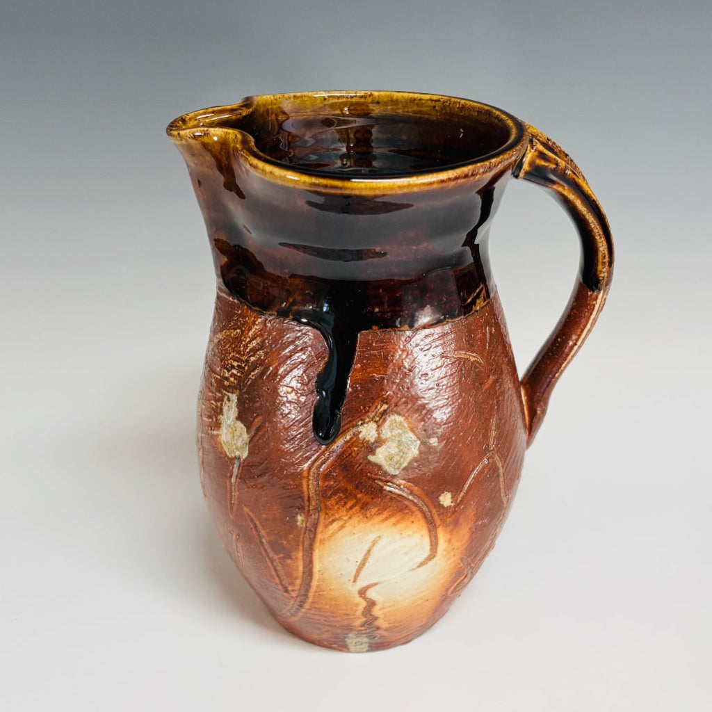 Handcrafted amber and chocolate brown wood-fired stoneware pitcher with pouring spout and handle.