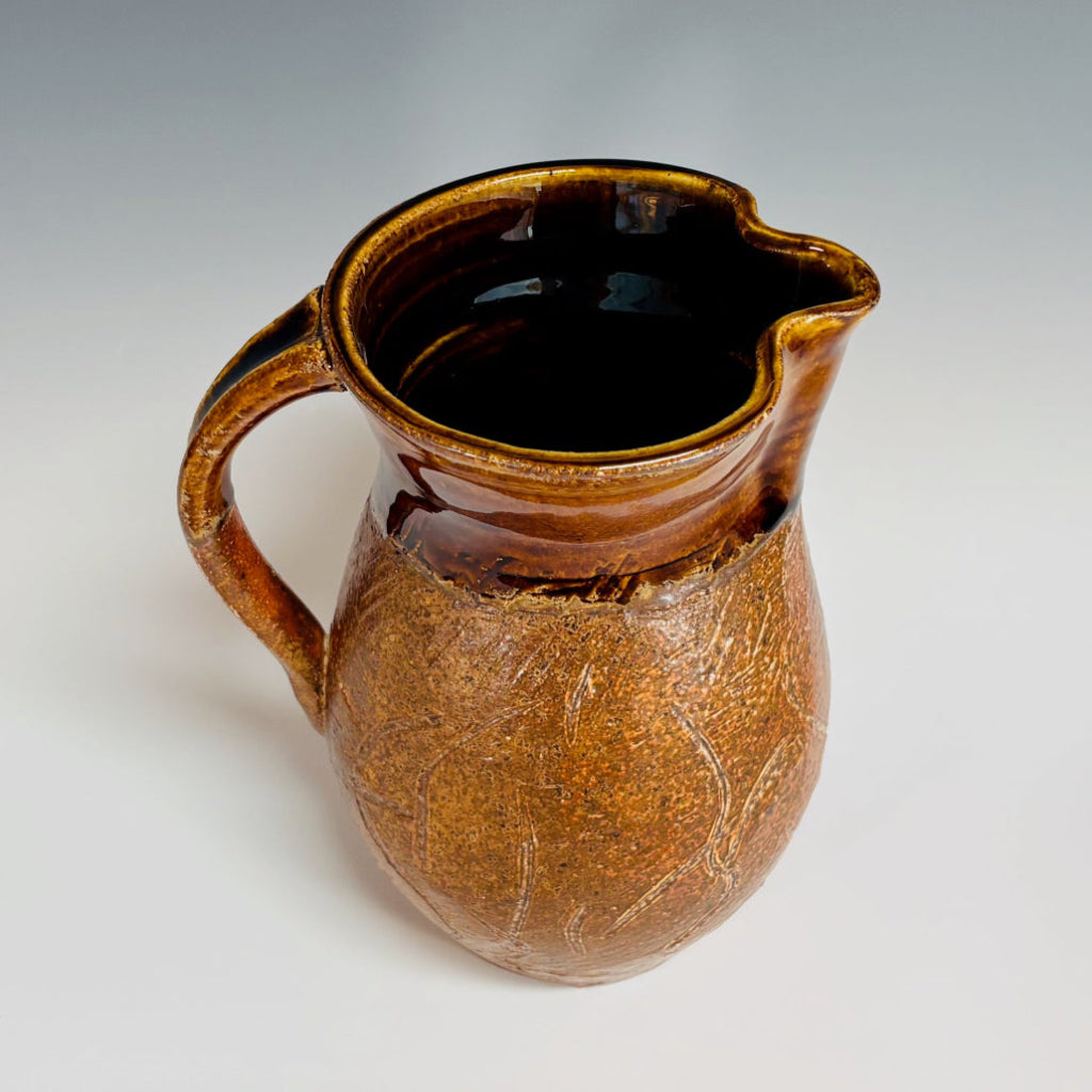 Side/Top view of hand-thrown stoneware pitcher showing glossy dark brown interior and textured surface and natural wood-firing effects.