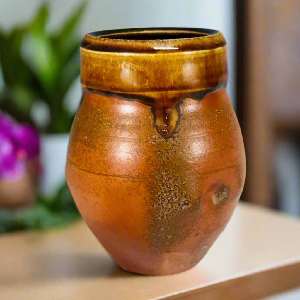 Rustic small vase with orange flashing  glaze on speckled rich amber clay, on a counter with flowers in the background. 