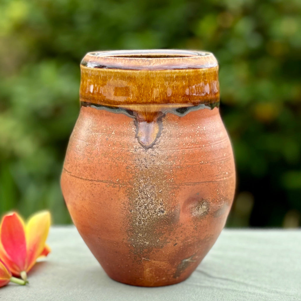 Rustic small vase with orange flashing  glaze on speckled rich amber clay, on a tabletop with a flower and greenery in the background. 