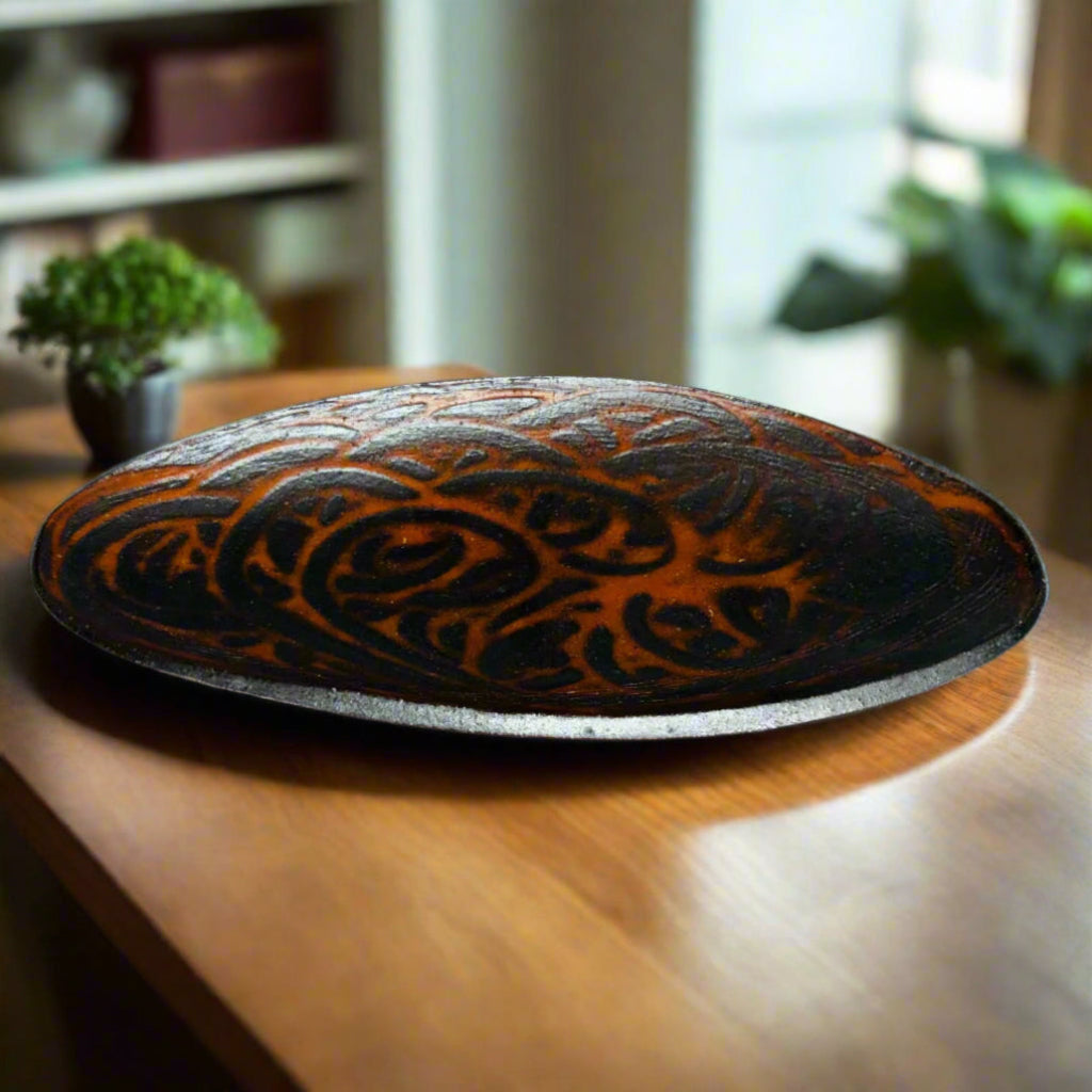 Decorative oval serving tray with delicate brown-black swirls over an orange flashing base on a kitchen countertop with plants and shelves in the background.