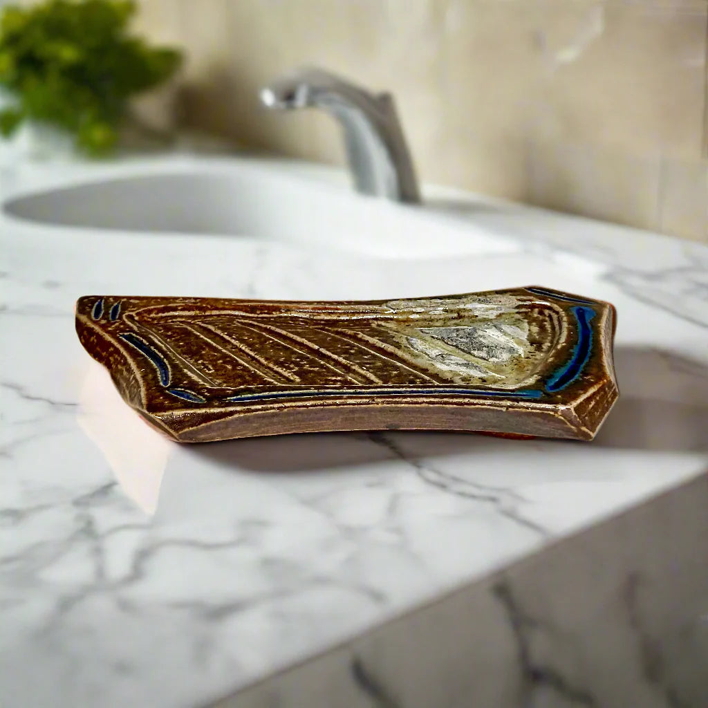 Handcrafted elongated soap dish with carved pattern and vibrant blue accents on brown stoneware sitting on marble sink top with a sink in the background.