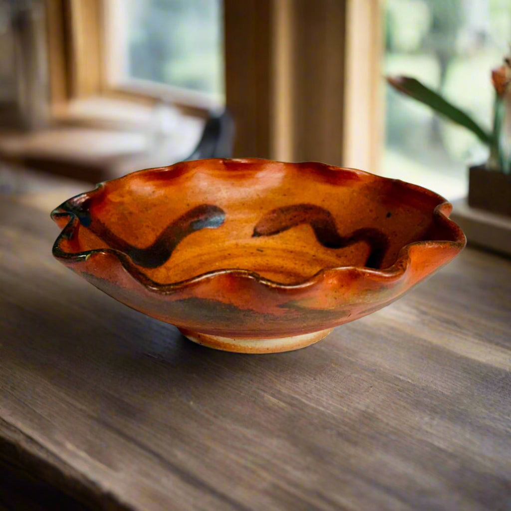 Handcrafted small ruffled ceramic bowl with amber-red wood-fired glaze and dark brushwork decoration on a wooden counter with window and plant in background, made in Danbury, NC.