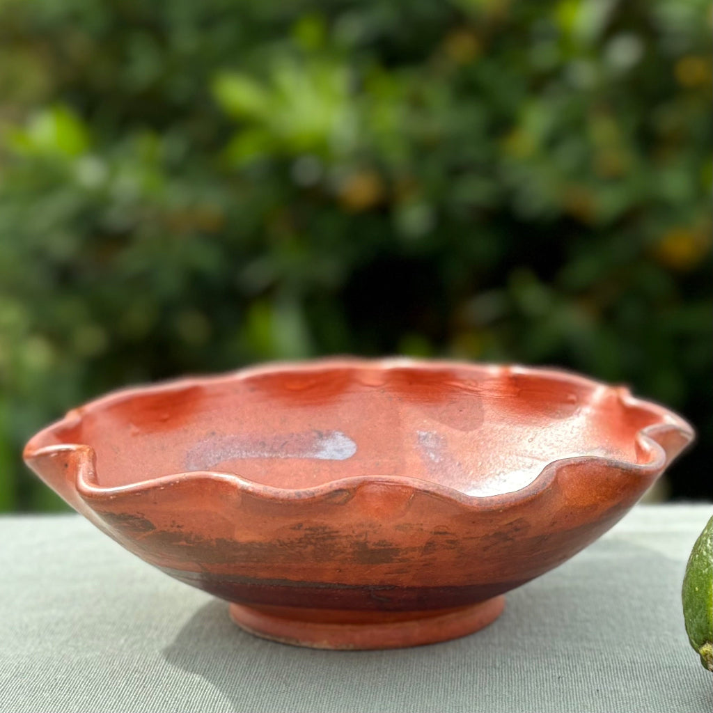 Handcrafted small ruffled ceramic bowl with amber-red wood-fired glaze and dark brushwork decoration on green cloth with greenery in the background, made in Danbury, NC.