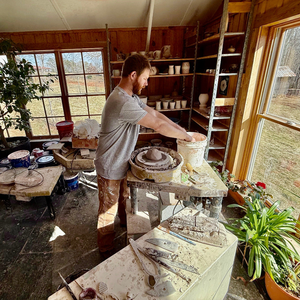 Sam Dougherty preparing to shape clay on the wheel into a pot with his hands over the edge of a water bucket.