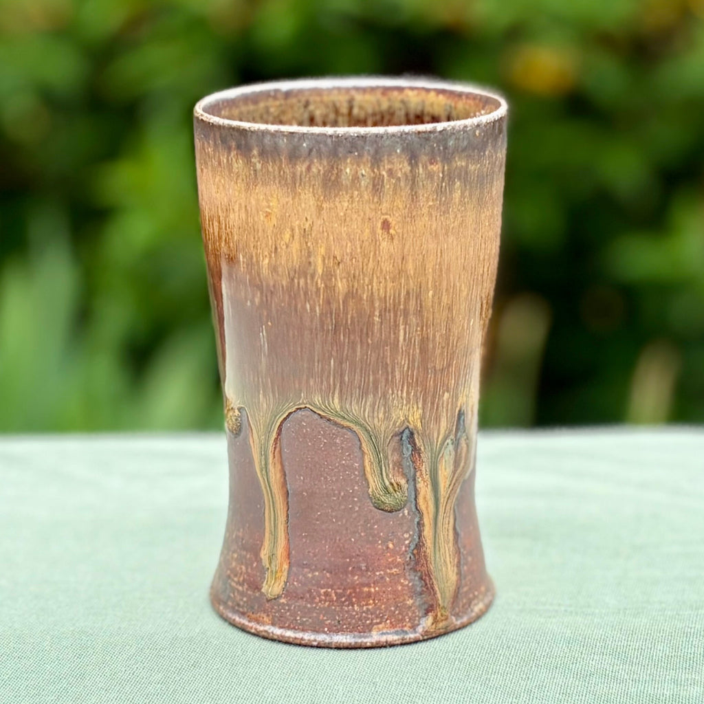 Hand-thrown stoneware tumbler with amber glaze drips on reddish-brown base from traditional wood-firing technique on a green cloth with greenery in the background. 