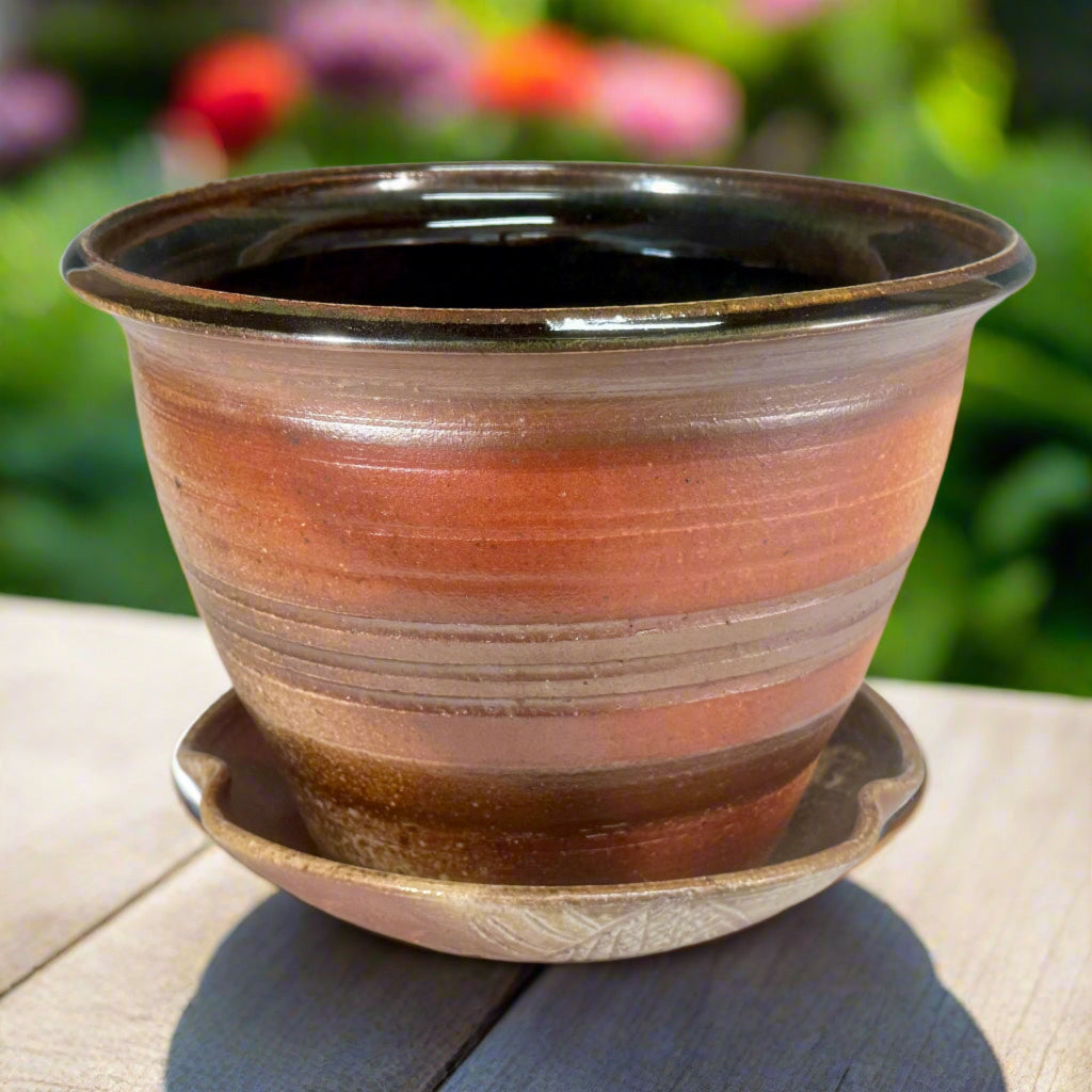 Artisan ceramic planter with amber and bronze glaze, hand-thrown in North Carolina with complementary drainage tray on table with flowers in the background. 