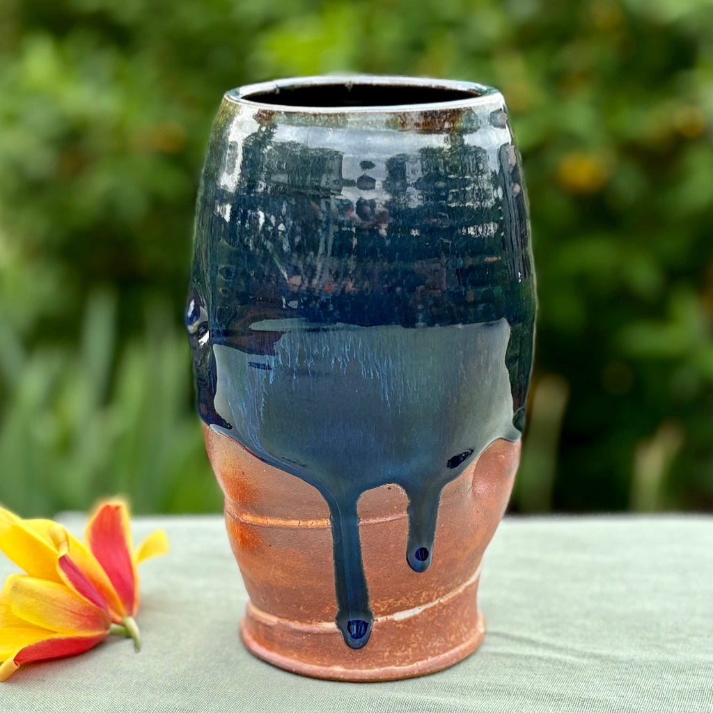 Handcrafted medium vase with cascading blue glaze over terracotta clay body set against of a living room, wood-fired in Danbury, NC on green cloth with flower and greenery in the background. 