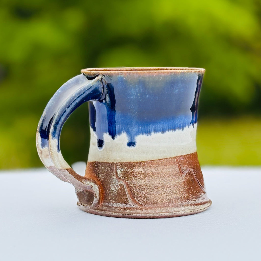 Side View Two: This pottery mug features a cobalt glaze rim dripping into a glossy white glaze with an unglazed orange at the bottom third of the mug. Delicate abstract decorations and walls that subtly taper outwards at the bottom. Made in Danbury, NC.
