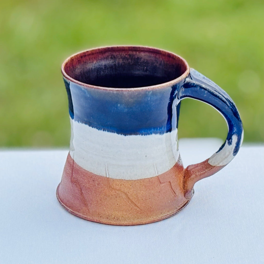 Top View: This pottery mug features a pink rim interior that fades to blue with a  cobalt glaze rim dripping into a glossy white glaze and unglazed orange at the bottom third on the exterior of the mug. Delicate abstract decorations and walls that subtly taper outwards at the bottom. Made in Danbury, NC.