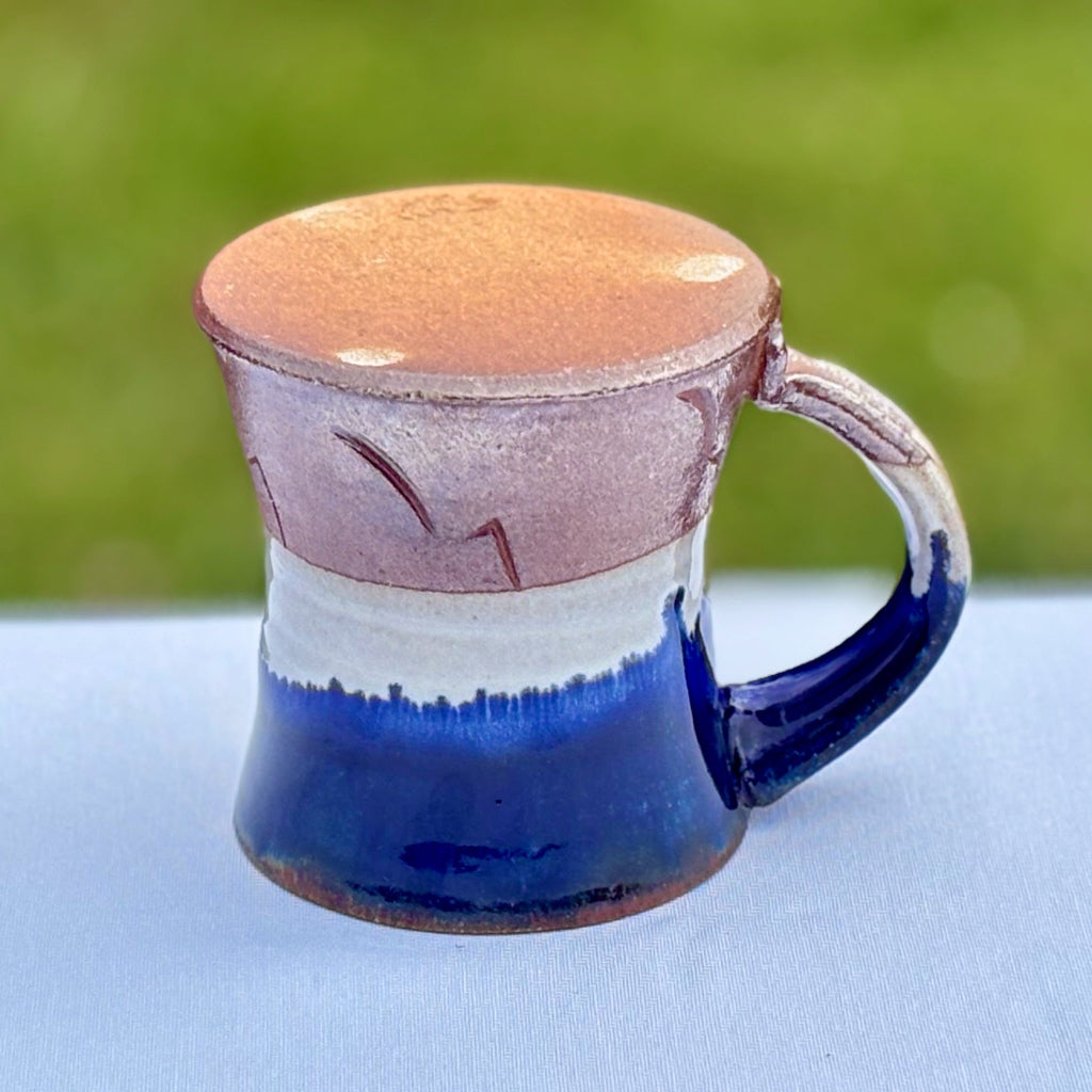 Bottom view: This pottery mug features an unglazed, warm amber bottom with artist's mark and a cobalt glaze rim dripping into a glossy white glaze and an unglazed orange at the bottom third of the mug. Delicate abstract decorations and  walls that subtly taper outwards at the bottom. Made in Danbury, NC.