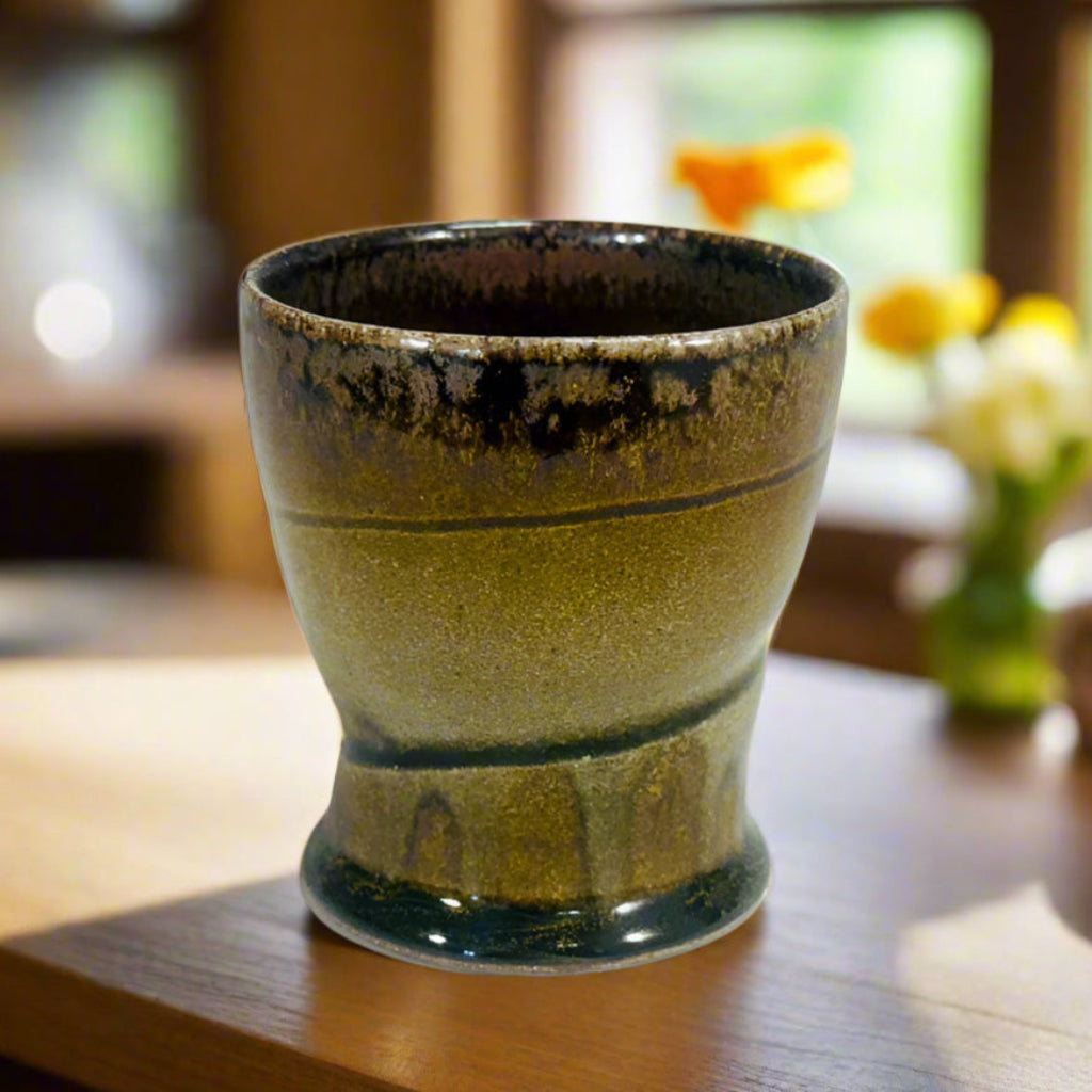 Handcrafted wood-fired ceramic tumbler with Handcrafted wood-fired ceramic tumbler with delicate greenish brown to chocolate transitions and horizontal banding on a counter with flowers and a window behind it. 