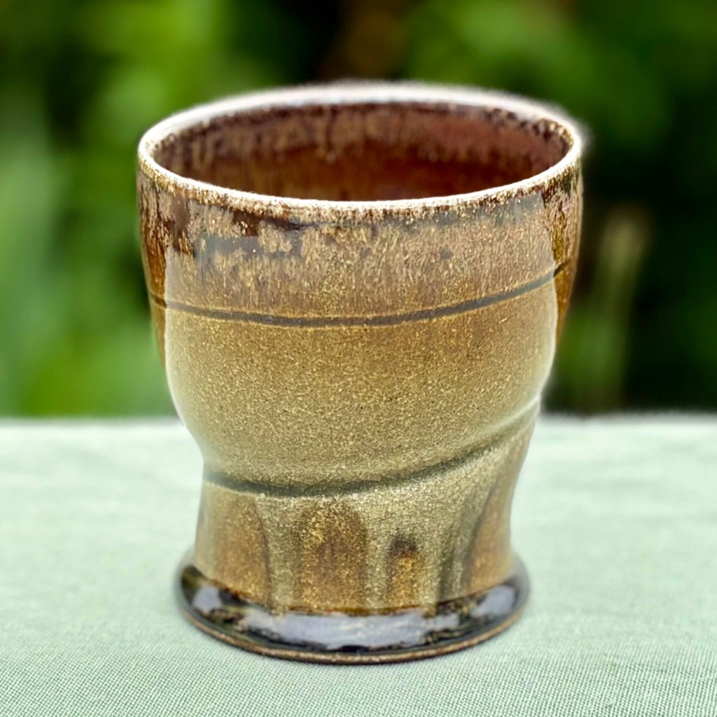 Handcrafted wood-fired ceramic tumbler with Handcrafted wood-fired ceramic tumbler with delicate greenish brown to chocolate transitions and horizontal banding on green tabletop with greenery in background. 