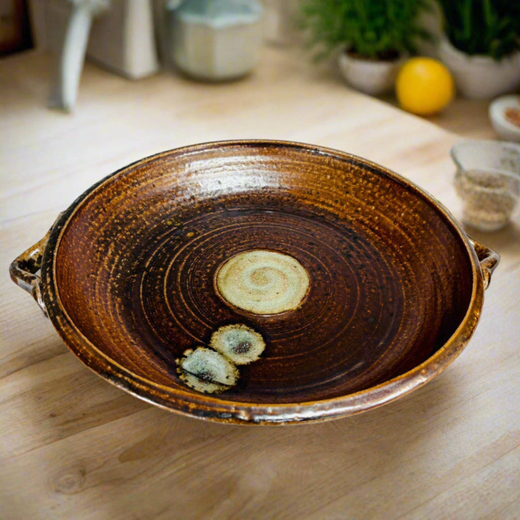  Handcrafted brown ceramic serving platter with integrated handles, concentric rings and cream spiral medallion center on a wooden countertop with plants and ingredients in the background.