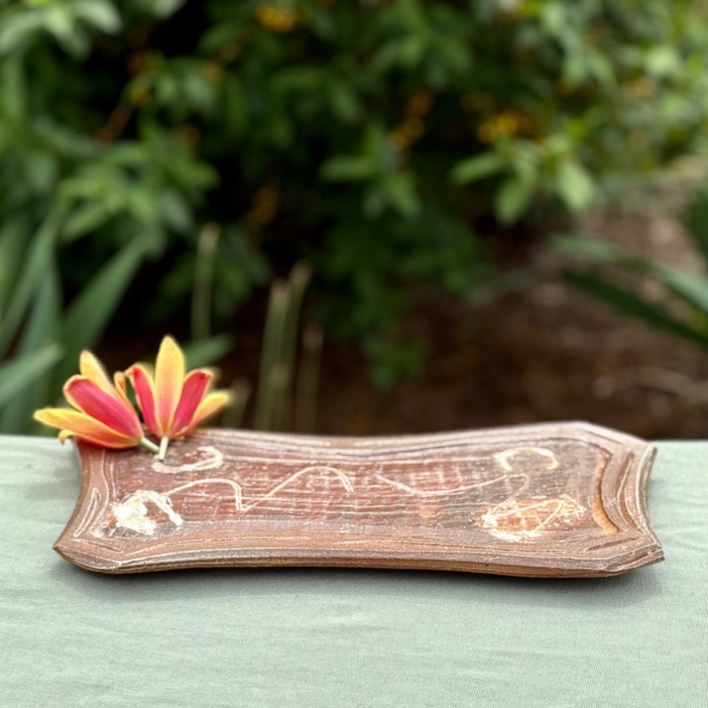 Handmade curved rectangular serving tray with white decorative accents on rich auburn surface, wood-fired in North Carolina on a green cloth and greenery in the background.