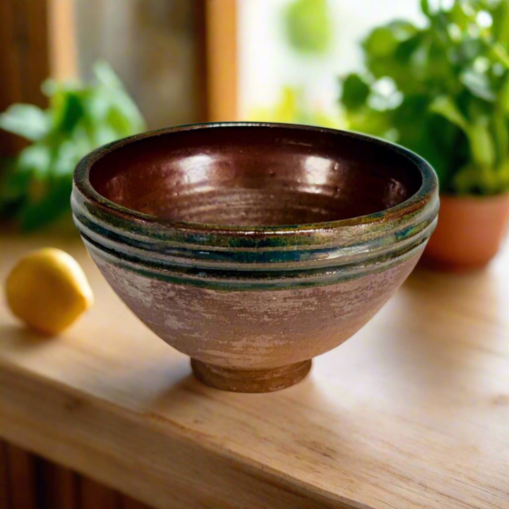 Handcrafted wood-fired bowl with teal green rim and earthy brown clay body, handmade in North Carolina on a wooden countertop with plants and a window in the background. 