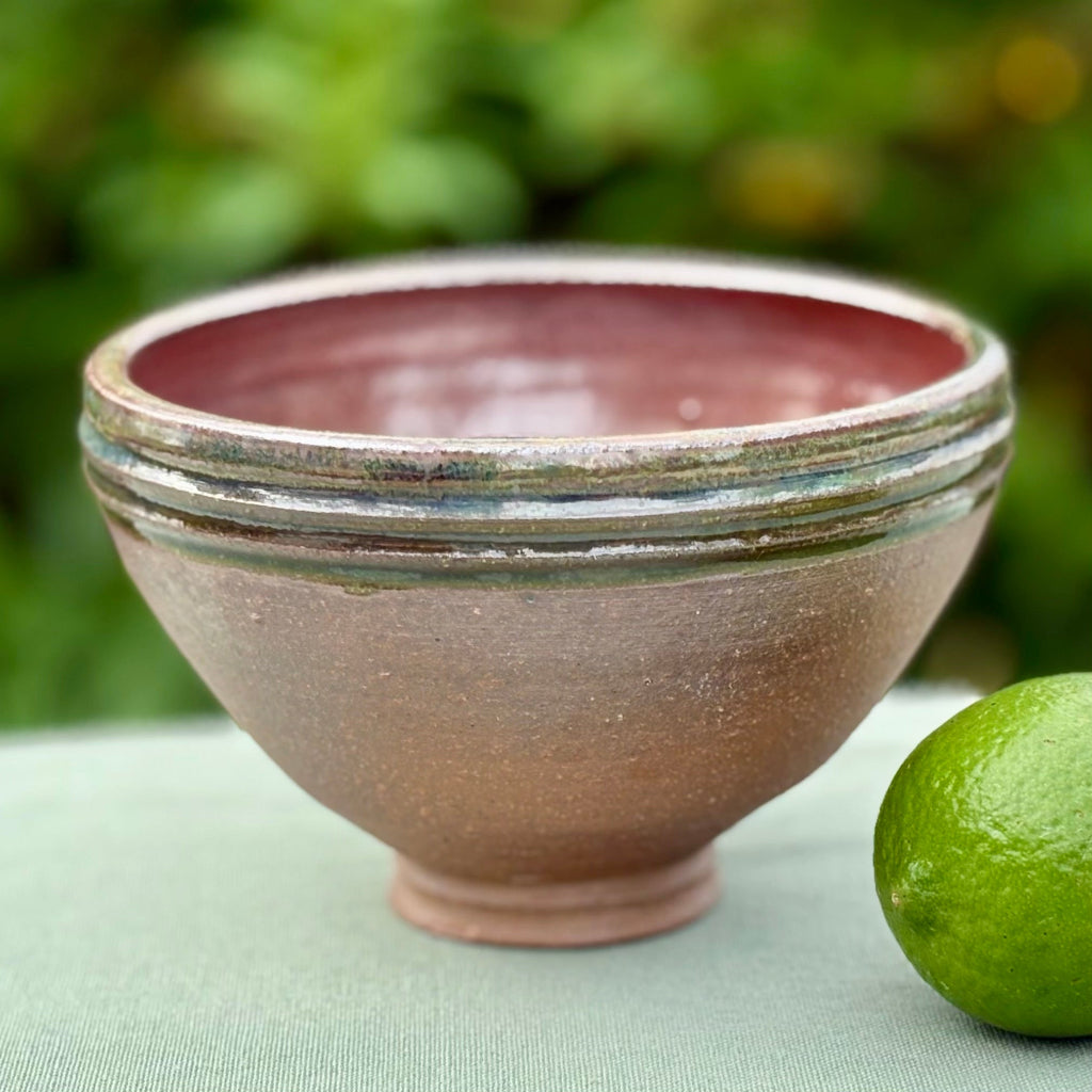 Pottery bowl with a green rim with three groves, brown interior and exterior and a delicate base on a green cloth with a lime and greenery in the background. 