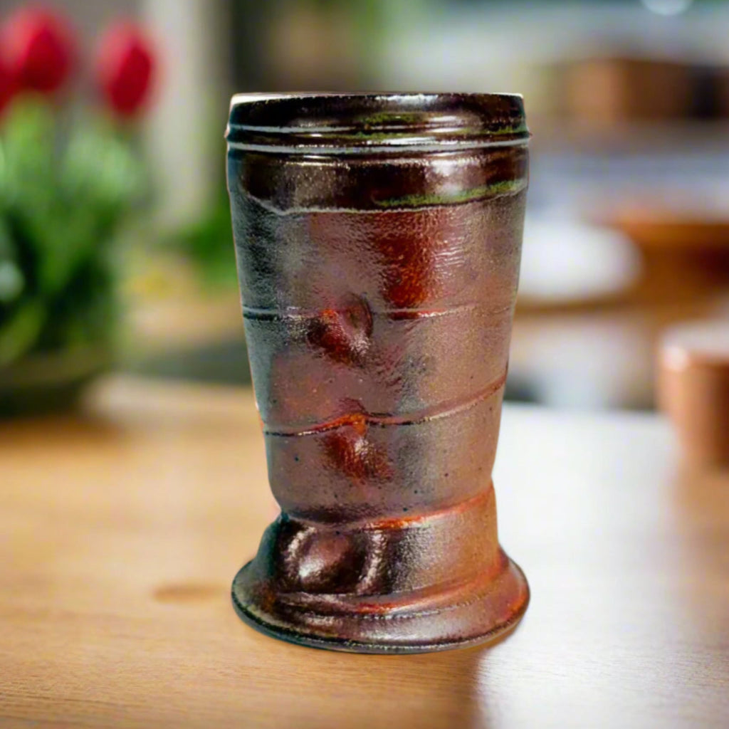 hand-thrown ceramic cup with horizontal ridges and a reddish-brown glaze, displayed on a table