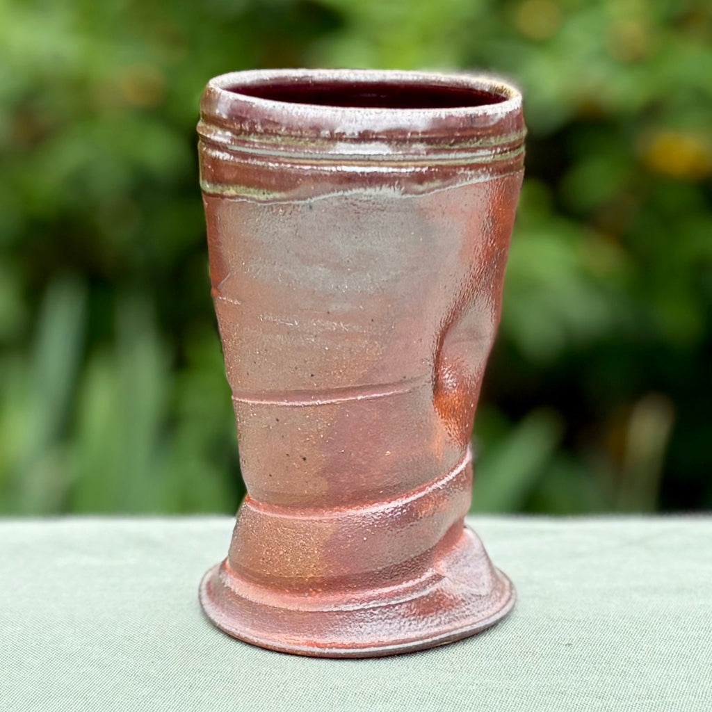 hand-thrown ceramic cup with horizontal ridges and a reddish-brown glaze, displayed against a blurred green background