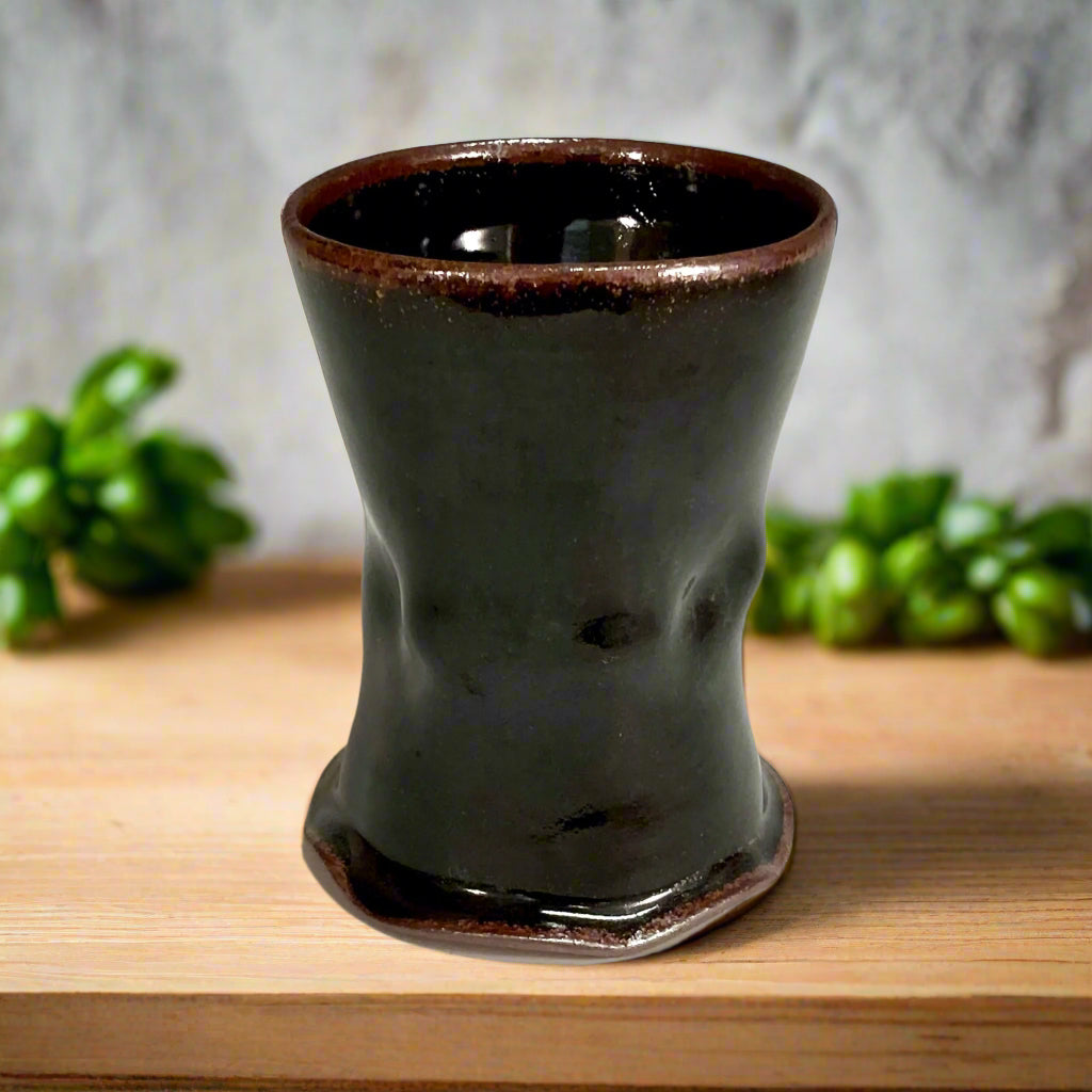 A stunning cup with Tenmoku glaze that creates a rich, shiny dark brown finish on a wooden countertop with basil in the background. 