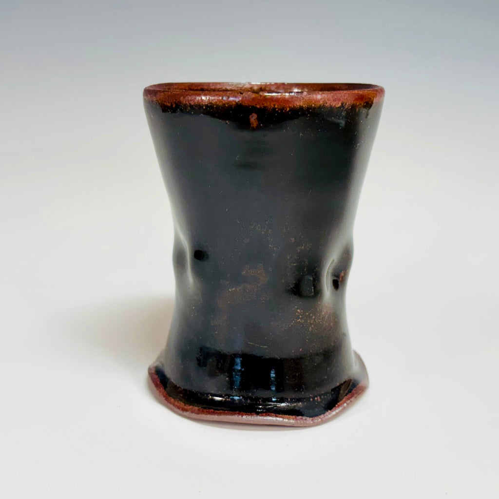 A stunning cup with Tenmoku glaze that creates a rich, shiny dark brown finish on a wooden countertop with basil in the background. 