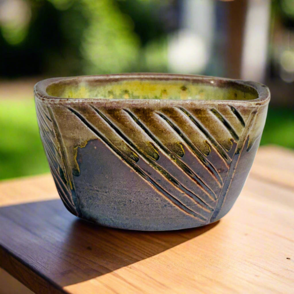 Purple-brown square ceramic planter with hand-carved diagonal pattern and yellow-green interior on a table in a yard.