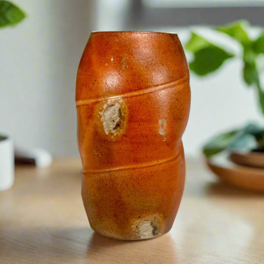 Handcrafted wood-fired ceramic vase in vibrant orange with natural ash deposits and horizontal banding on wooden countertop with window and plants behind window.