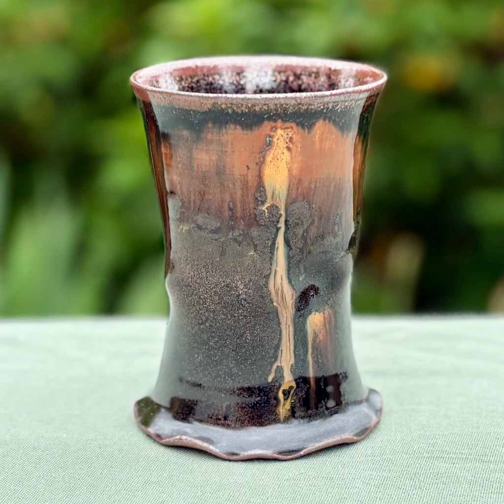This cup is a handcrafted wood-fired cup with dark brown/black glaze with amber drip from rim to base, and speckled rim on green cloth with greenery in the background. 
