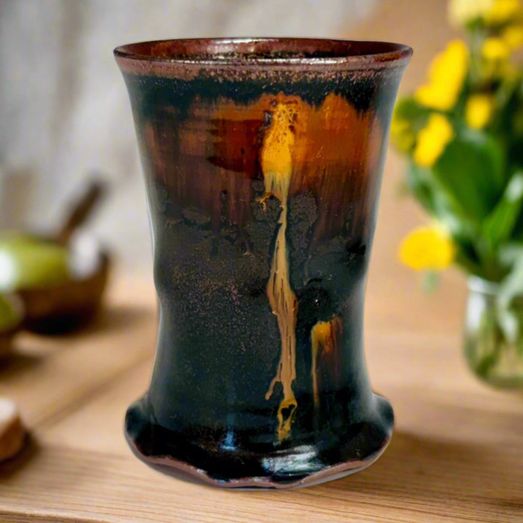 This cup is a handcrafted wood-fired cup with dark brown/black glaze with amber drip from rim to base, and speckled rim on a wooden countertop.
