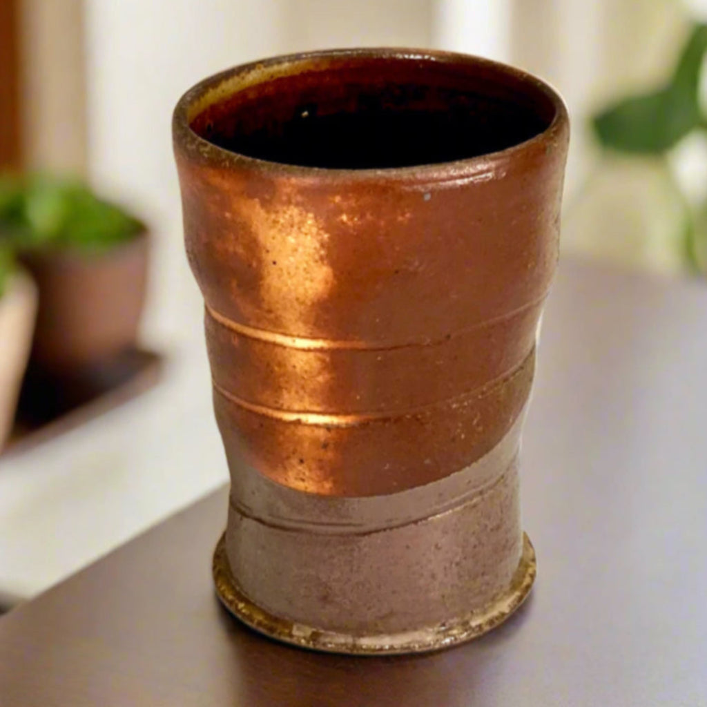 Hand-thrown copper-banded tenmoku cup with distinctive ridges, wood-fired in historic NC kiln sitting on a wooden table with plants in the background.
