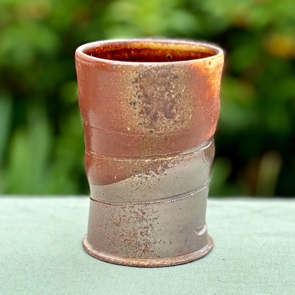 Hand-thrown copper-banded tenmoku cup with distinctive ridges, wood-fired in historic NC kiln sitting on a green cloth with greenery in the background. 