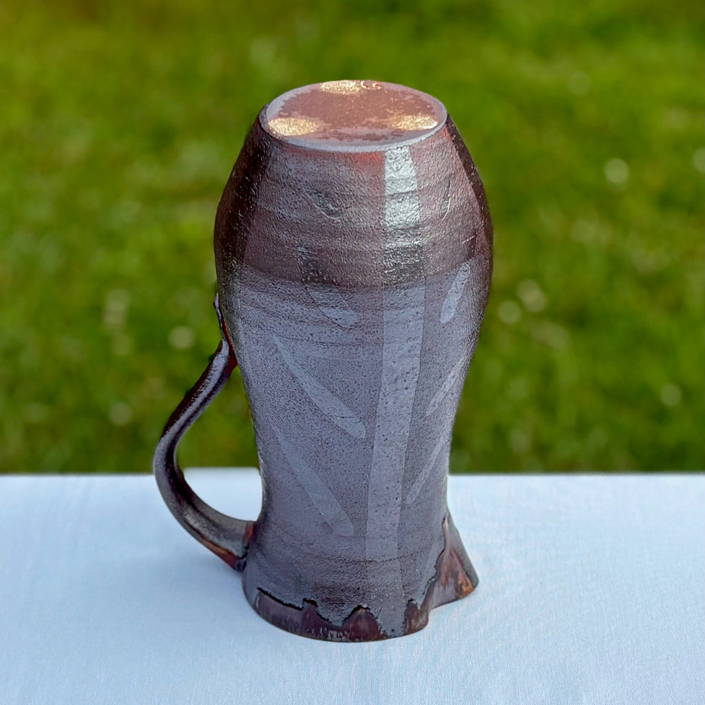 Bottom view: Unglazed pottery pitche rbottom with three white dots and an artist's mark. A dark tenmoku glaze contrasts with an orange and brown flashing resulting from firing in a wood kiln. Lovely drip rim with vibrant glossy orange bleeding into darker browns. A subtle pattern of abstract vertical and diagonal lines add visual interest to the outside of this pot. Made in Danbury, NC. 