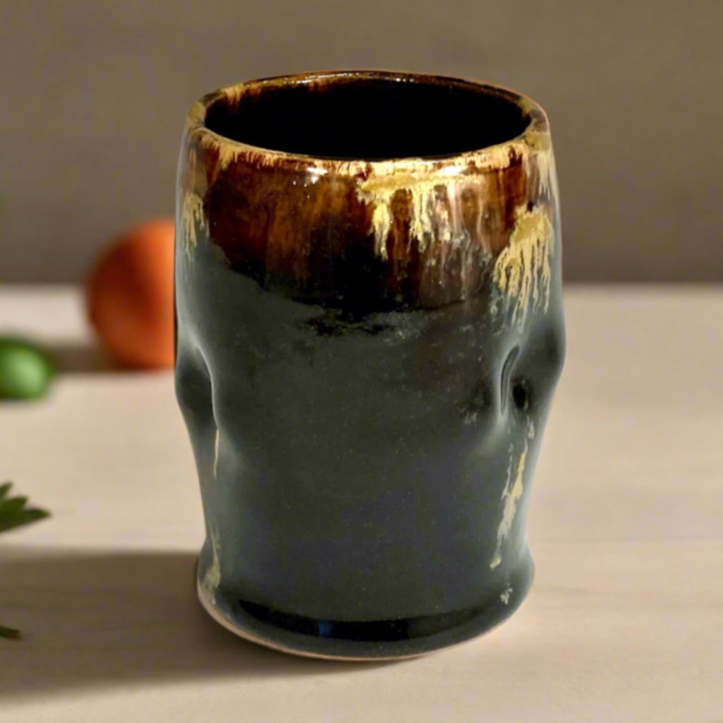 Handcrafted black stoneware tumbler with golden amber drip glaze from wood-fired kiln on countertop with fruit in the background. 