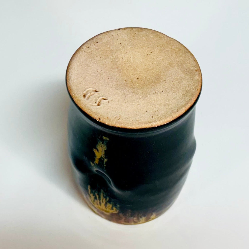 Bottom view: Handcrafted black stoneware tumbler with golden amber drip glaze from wood-fired kiln.