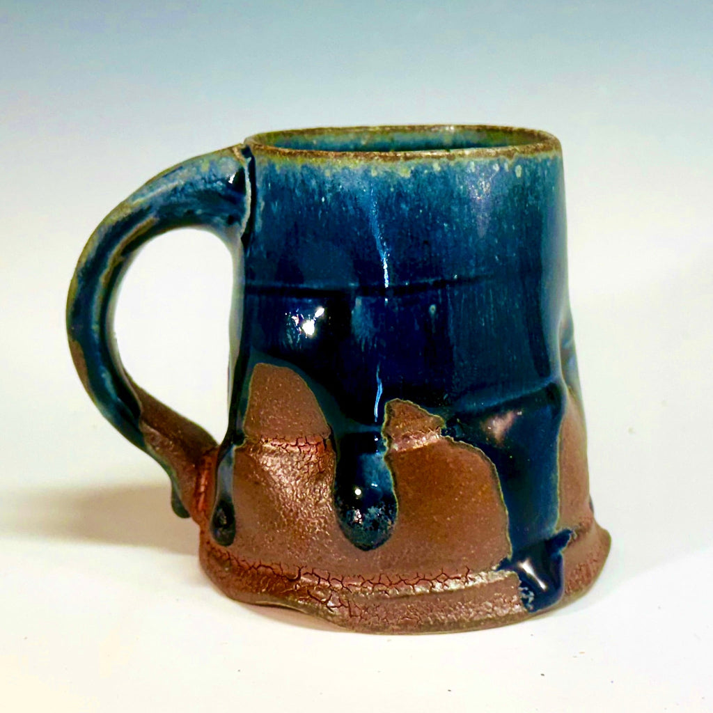 Handcrafted wood-fired ceramic mug with dramatic midnight blue glaze dripping over earthy clay body and sturdy handle.