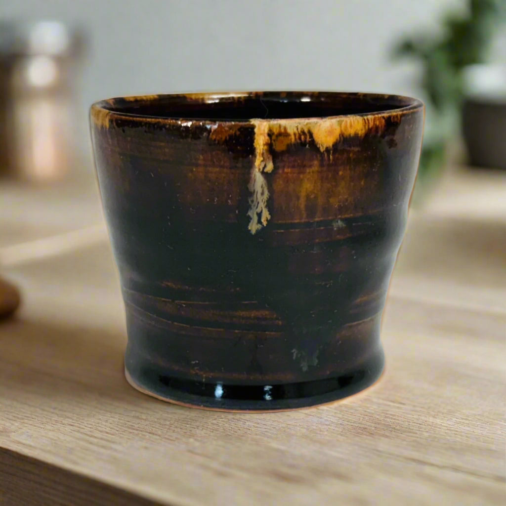 Handmade ceramic cup with dark tenmoku glaze and amber drip detail, wheel-thrown pottery cup, artisanal stoneware vessel with glossy finish on a kitchen countertop.