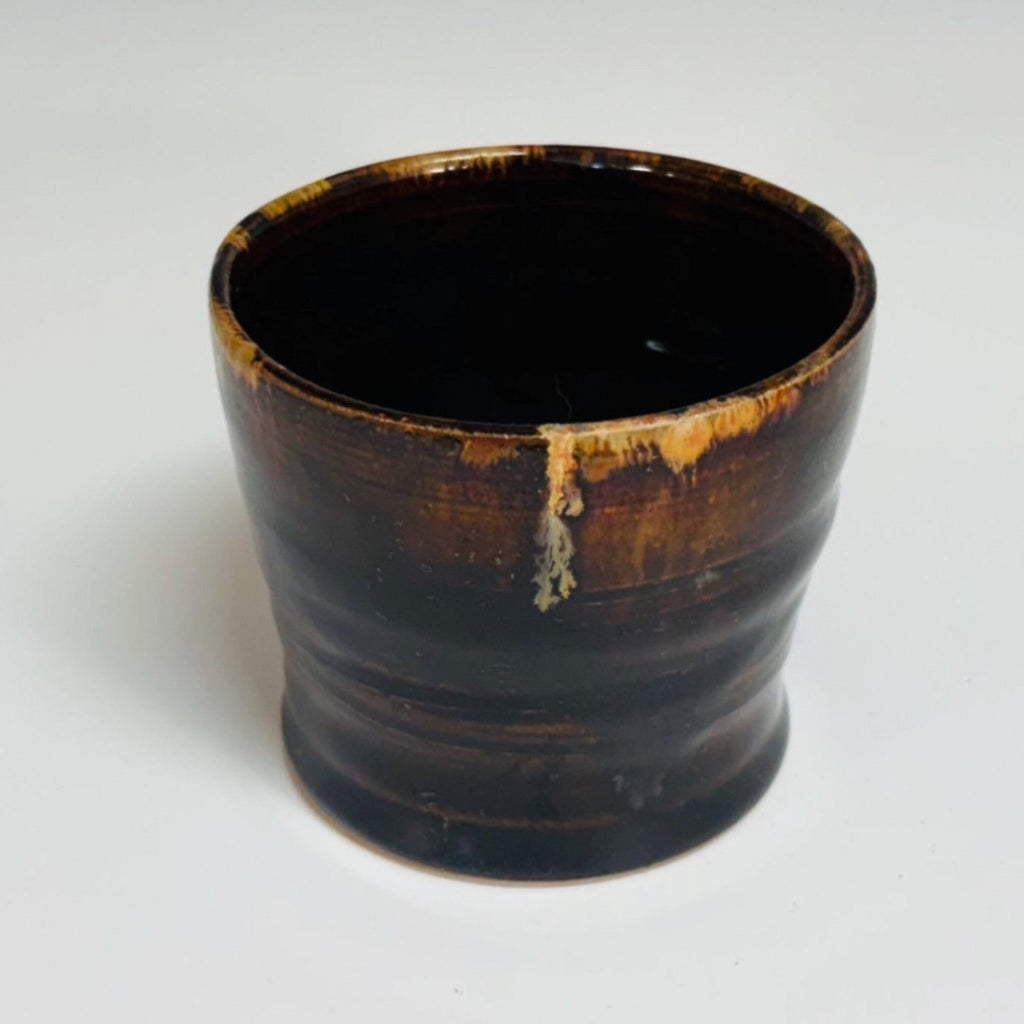 Handmade ceramic cup with dark tenmoku glaze and amber drip detail, wheel-thrown pottery cup, artisanal stoneware vessel with glossy finish.