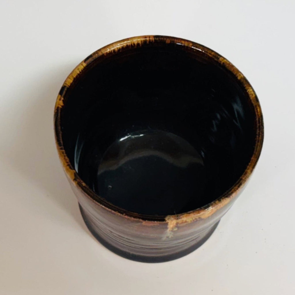 Handmade ceramic cup with dark tenmoku glaze and amber drip detail, wheel-thrown pottery cup, artisanal pottery vessel with glossy finish.